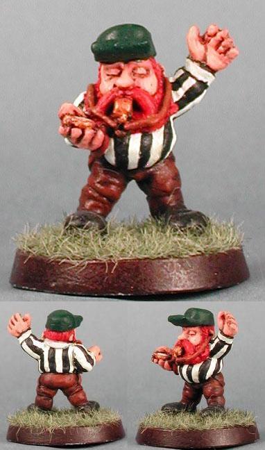 Painted miniature of a red-bearded Dwarf referee in a striped shirt on a grassy base.