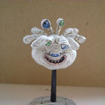 White clay face sculpture on a black stand, with blue eyes and a wide grin.